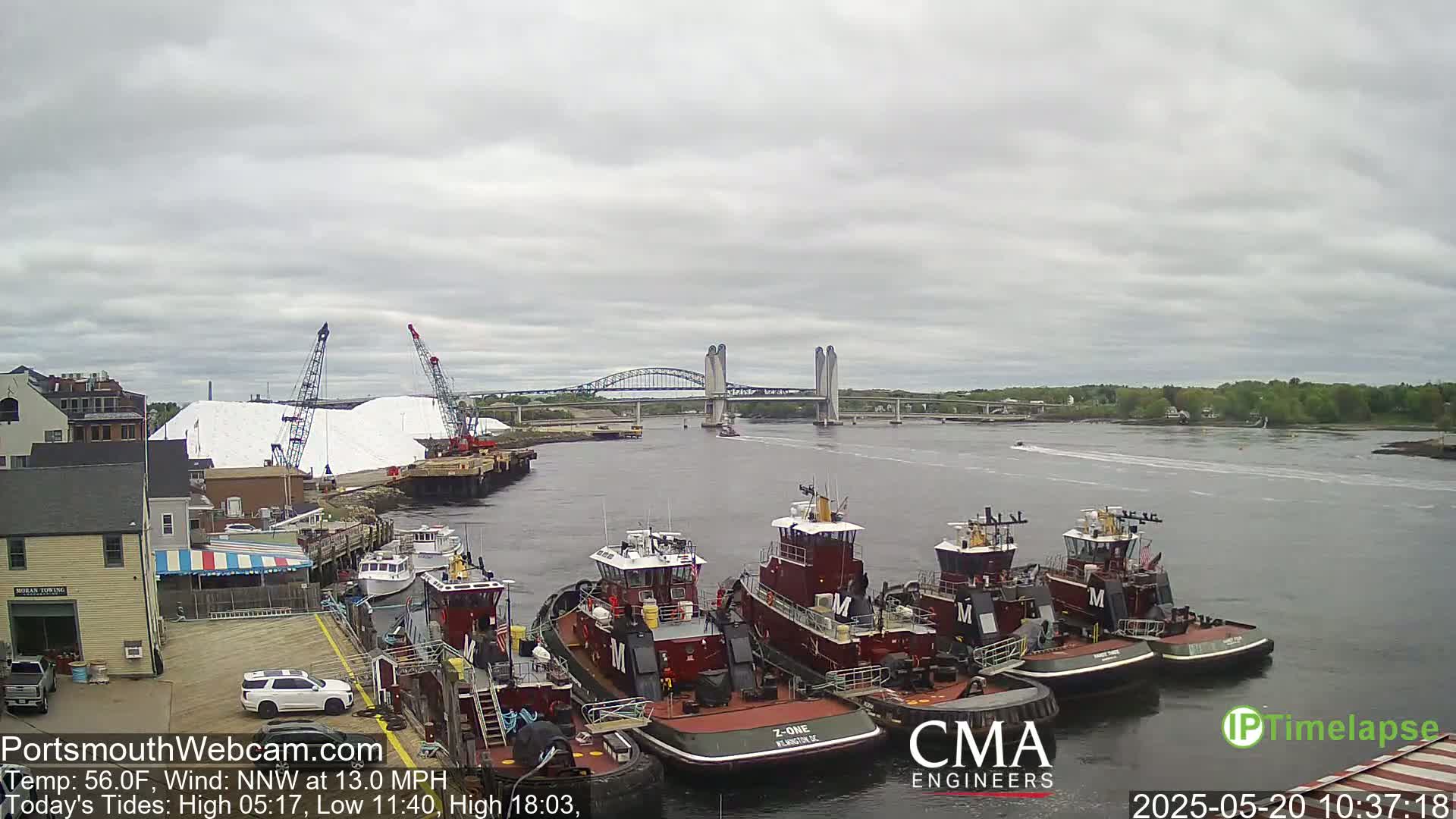 Several tugboats are moored at a dock near a bridge under an overcast sky, with a large pile of white material and cranes visible in the background.