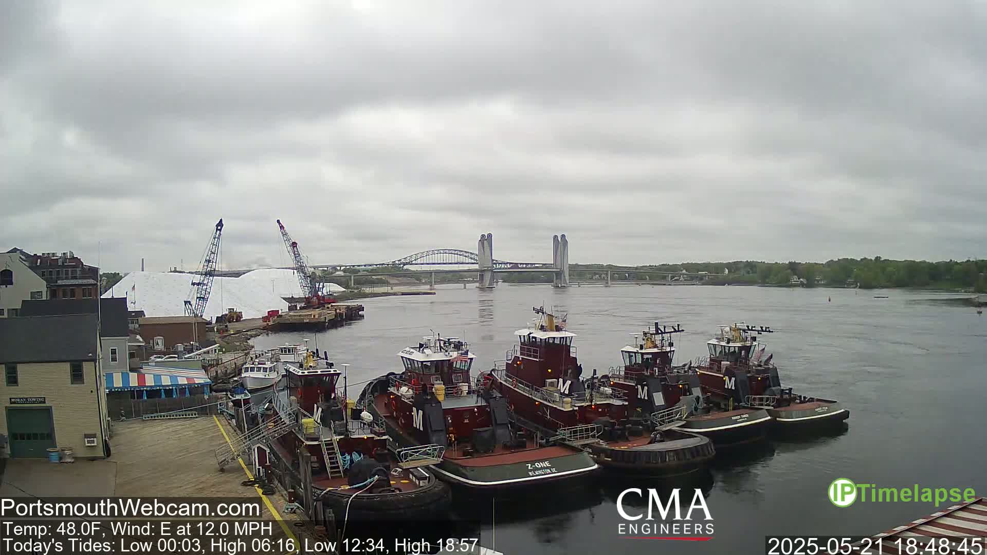 Several tugboats are moored at a dock next to a large pile of white material, with a bridge spanning a river in the background under an overcast sky.