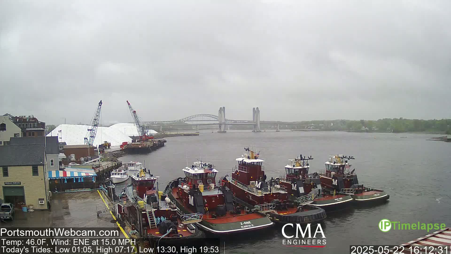 Several tugboats are moored at a dock next to large piles of white material, under an overcast sky with a moderate wind, in front of a lift bridge spanning a waterway.