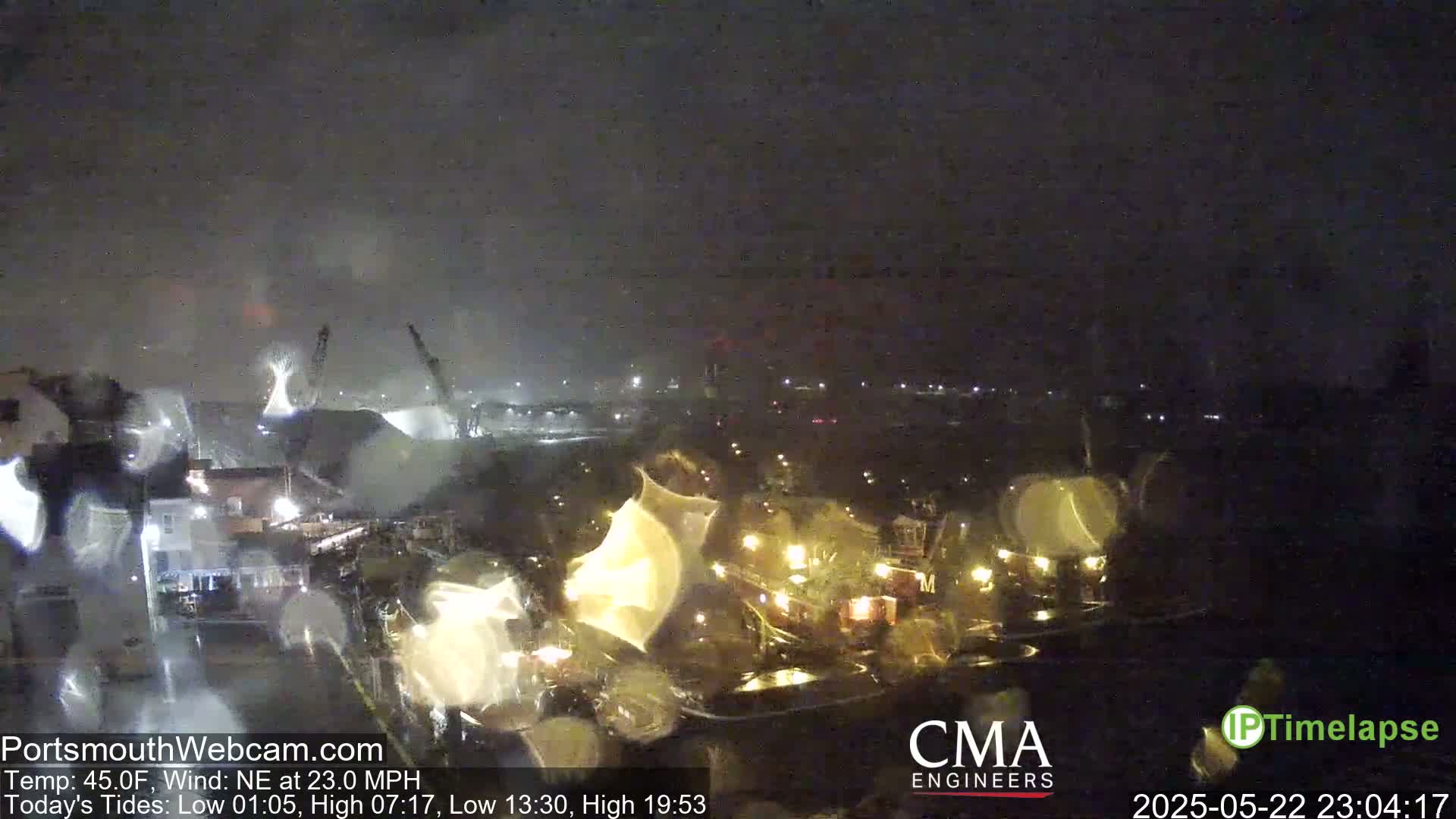 A nighttime view of a harbor with several illuminated boats and buildings under a dark, rainy sky with a strong northeast wind.