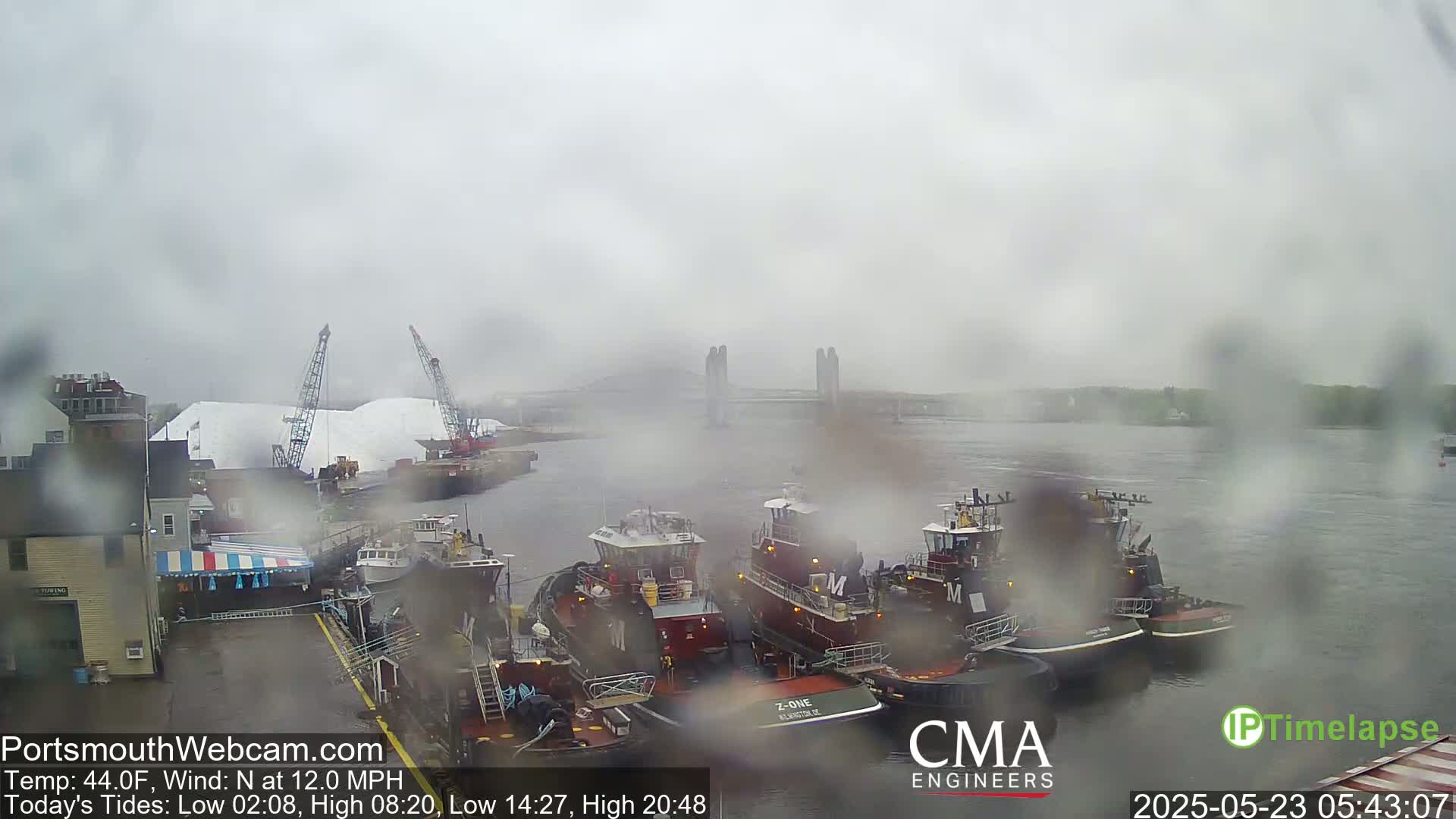 Several tugboats are moored at a dock in rainy conditions, with a large pile of white material and cranes visible in the background across a waterway.