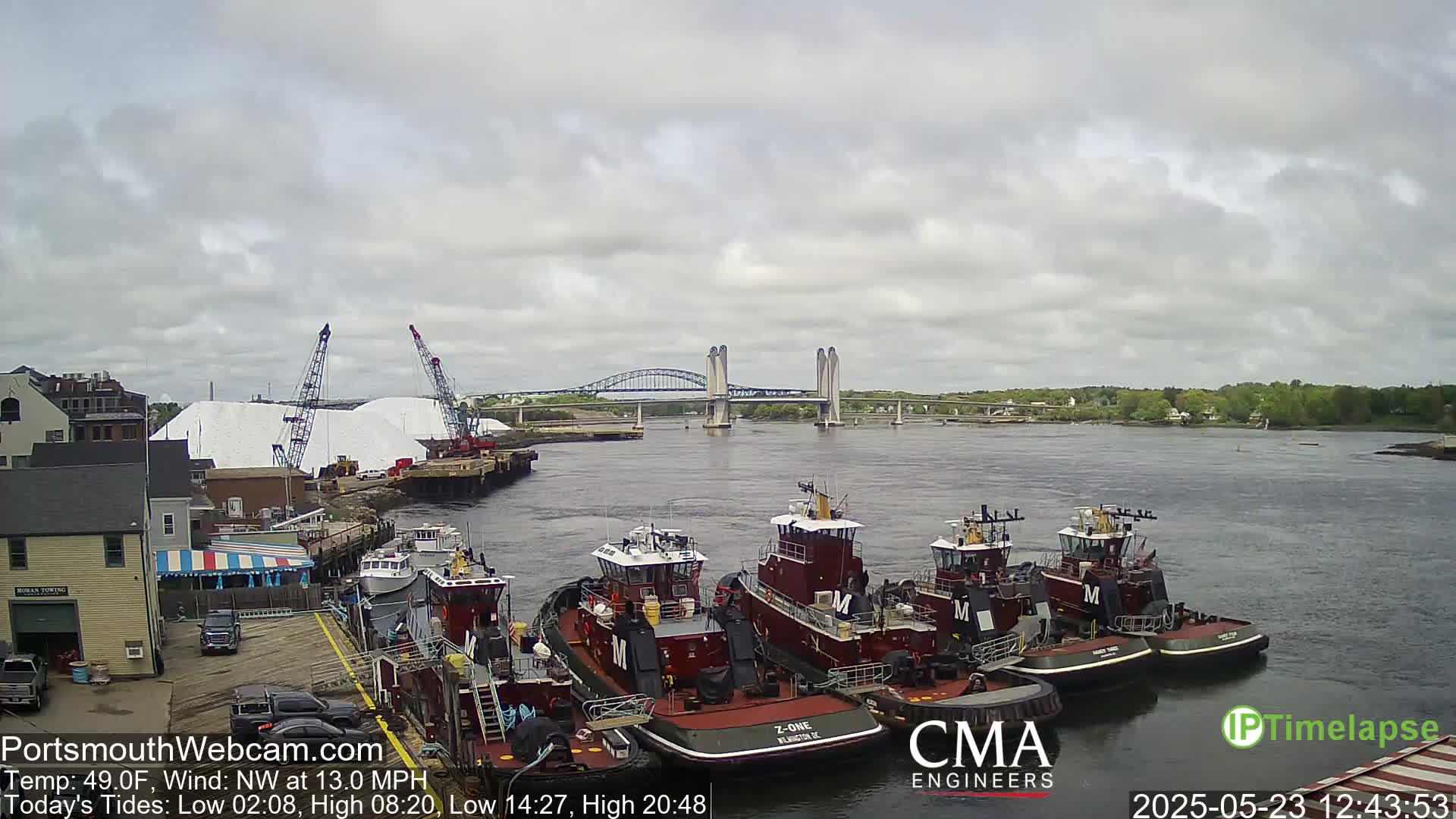 Several tugboats are moored at a dock next to piles of white material, with a bridge spanning a river in the background under a cloudy sky.