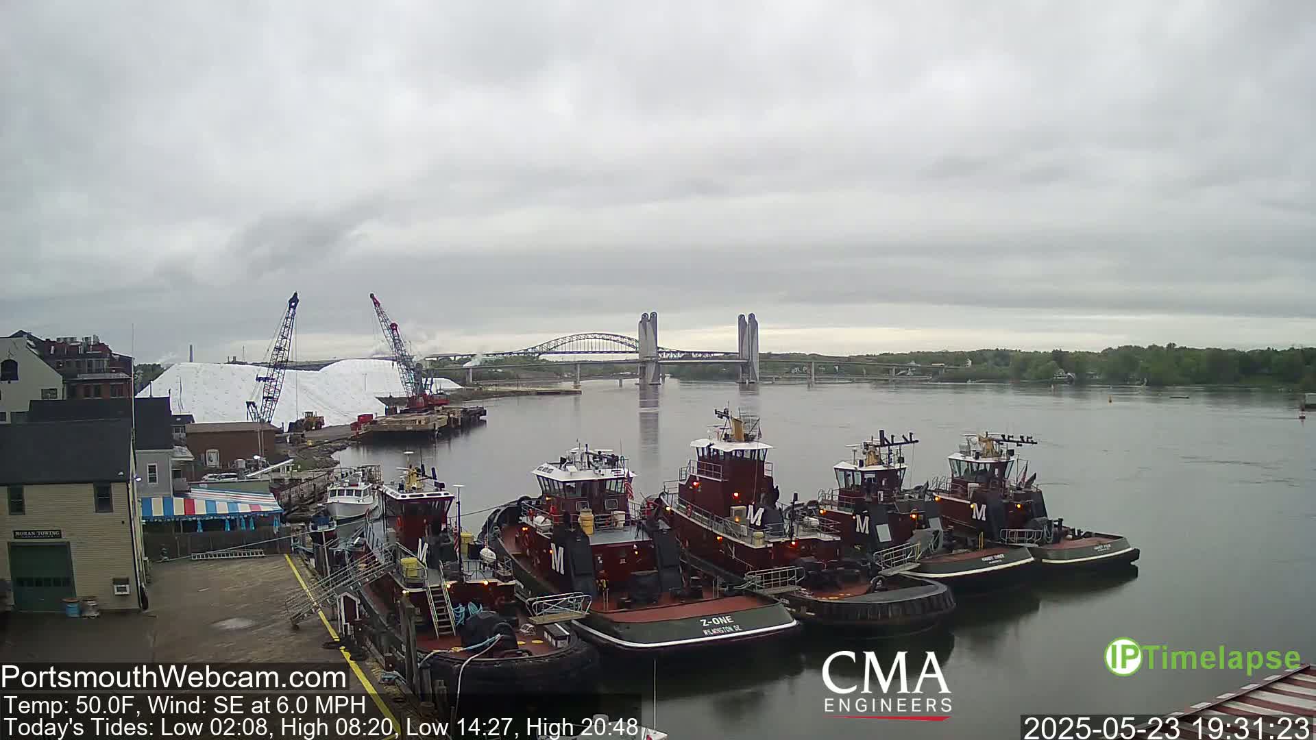 Several tugboats are moored at a dock next to a large pile of white material, with a bridge spanning a river in the background under an overcast sky.