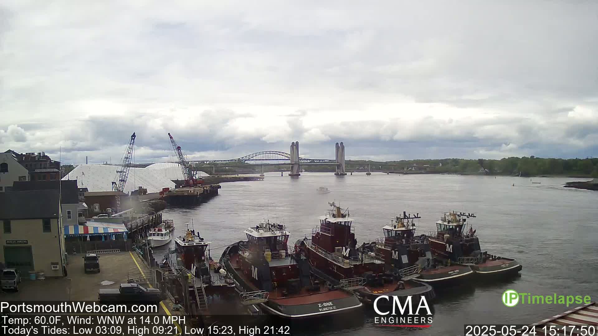 Several tugboats are moored at a dock next to a large pile of salt, with a lift-bridge spanning a river under a cloudy sky.