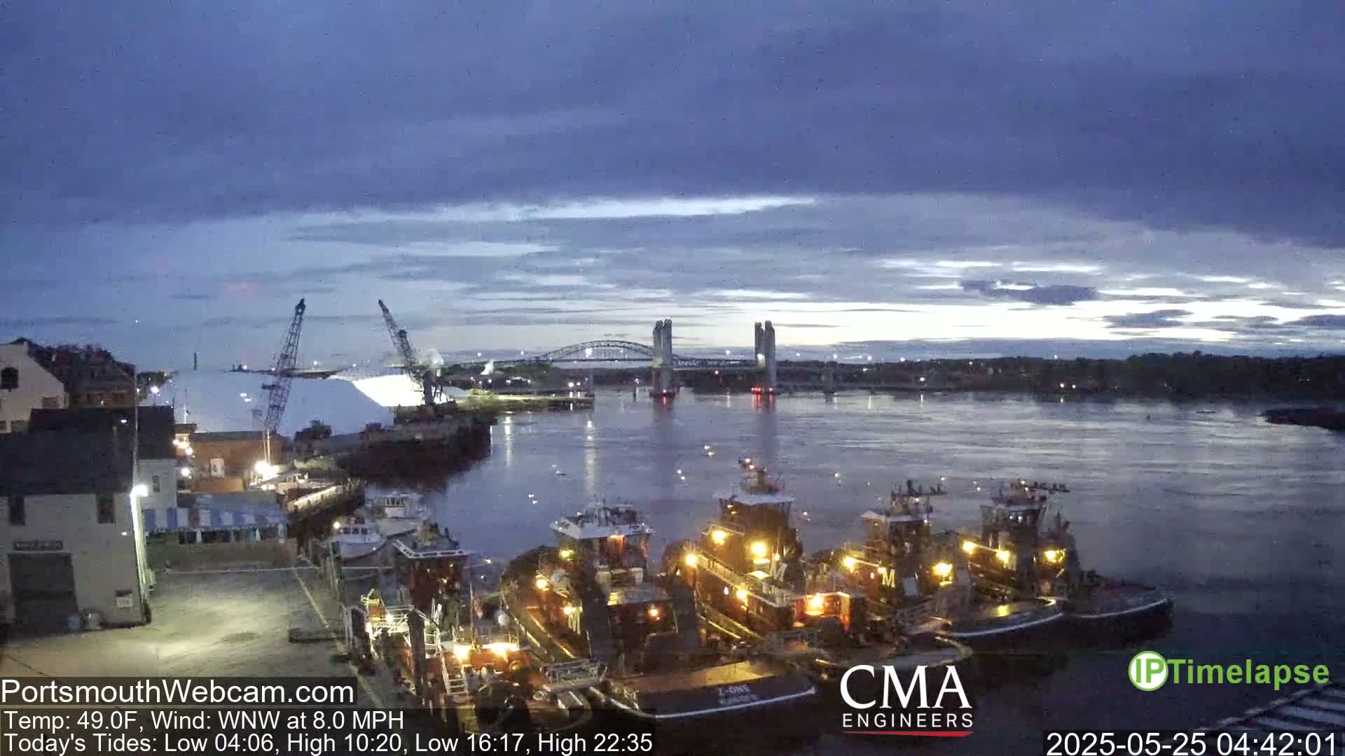 Several tugboats are docked at a harbor under a twilight sky with a bridge in the distance, and a large pile of white material is visible on the left.