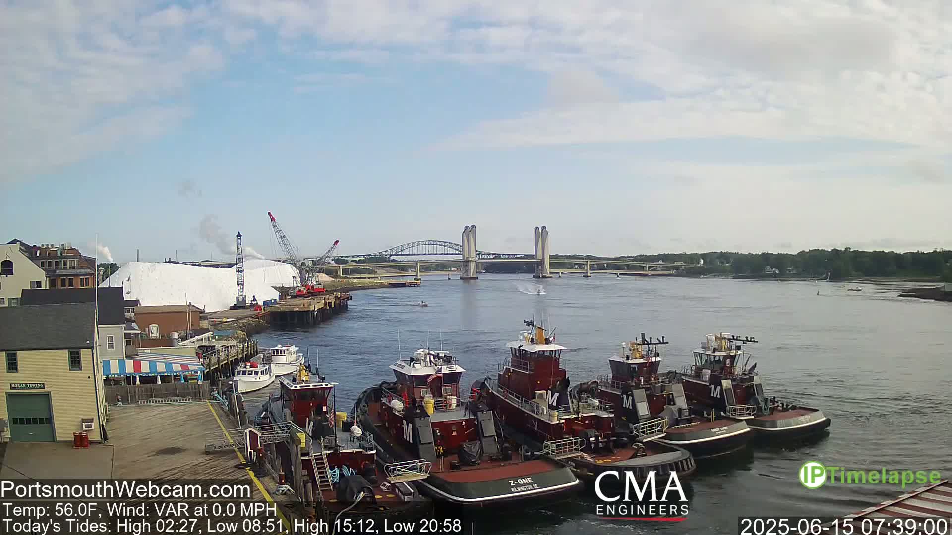Several tugboats are moored at a dock next to a large pile of white material, with a bridge and calm water in the background under a partly cloudy sky.