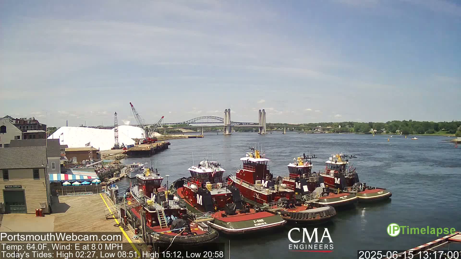 Several tugboats are docked at a waterfront, with a large pile of white material and a bridge in the background on a sunny day with an east wind.