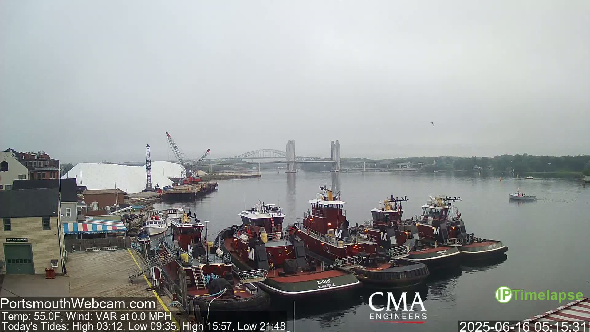 Several tugboats are moored at a dock near a bridge on a foggy day, with a large pile of white material in the background.