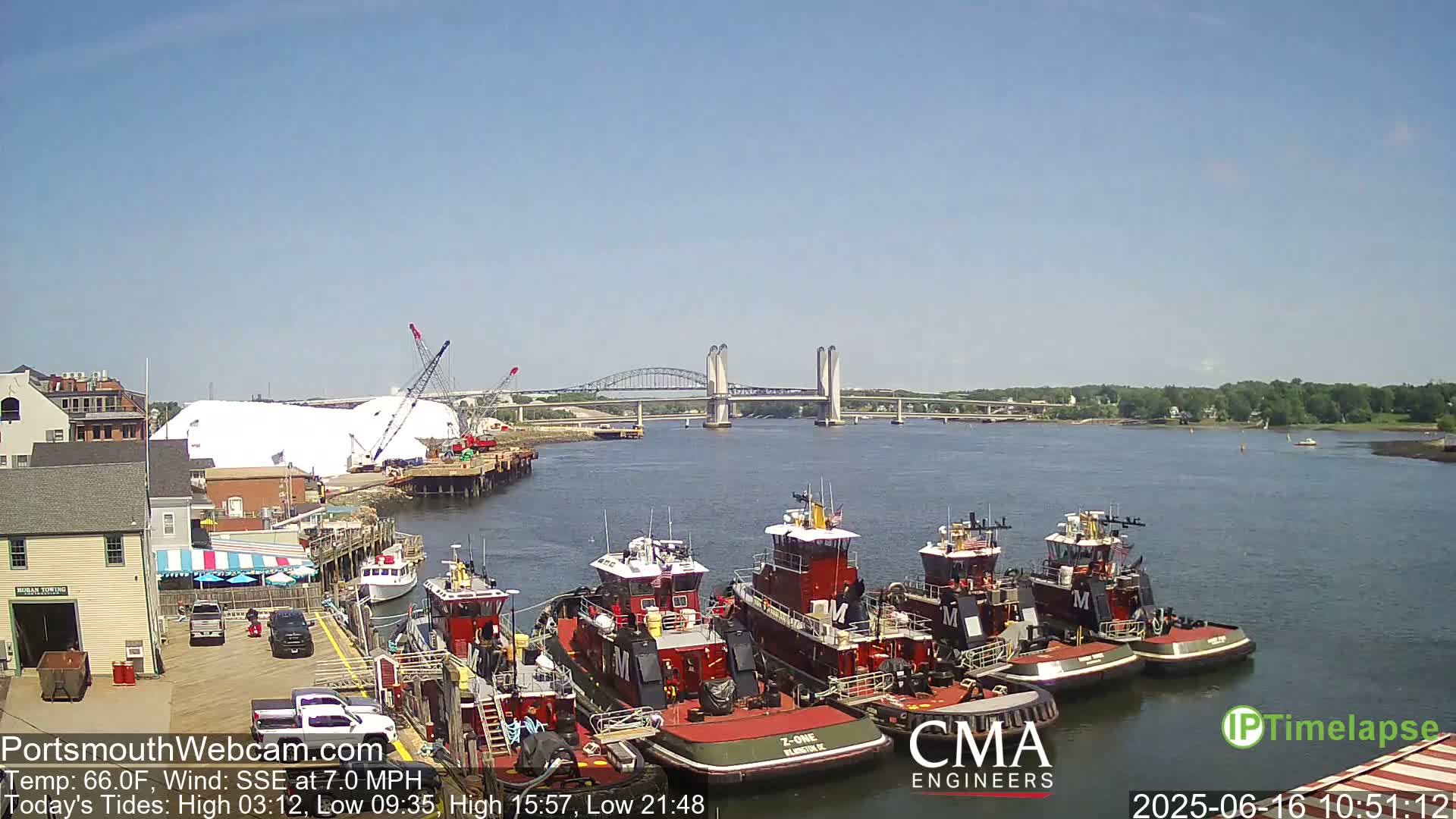Several tugboats are moored at a waterfront dock, with a large pile of material and a bridge visible in the background on a sunny day.