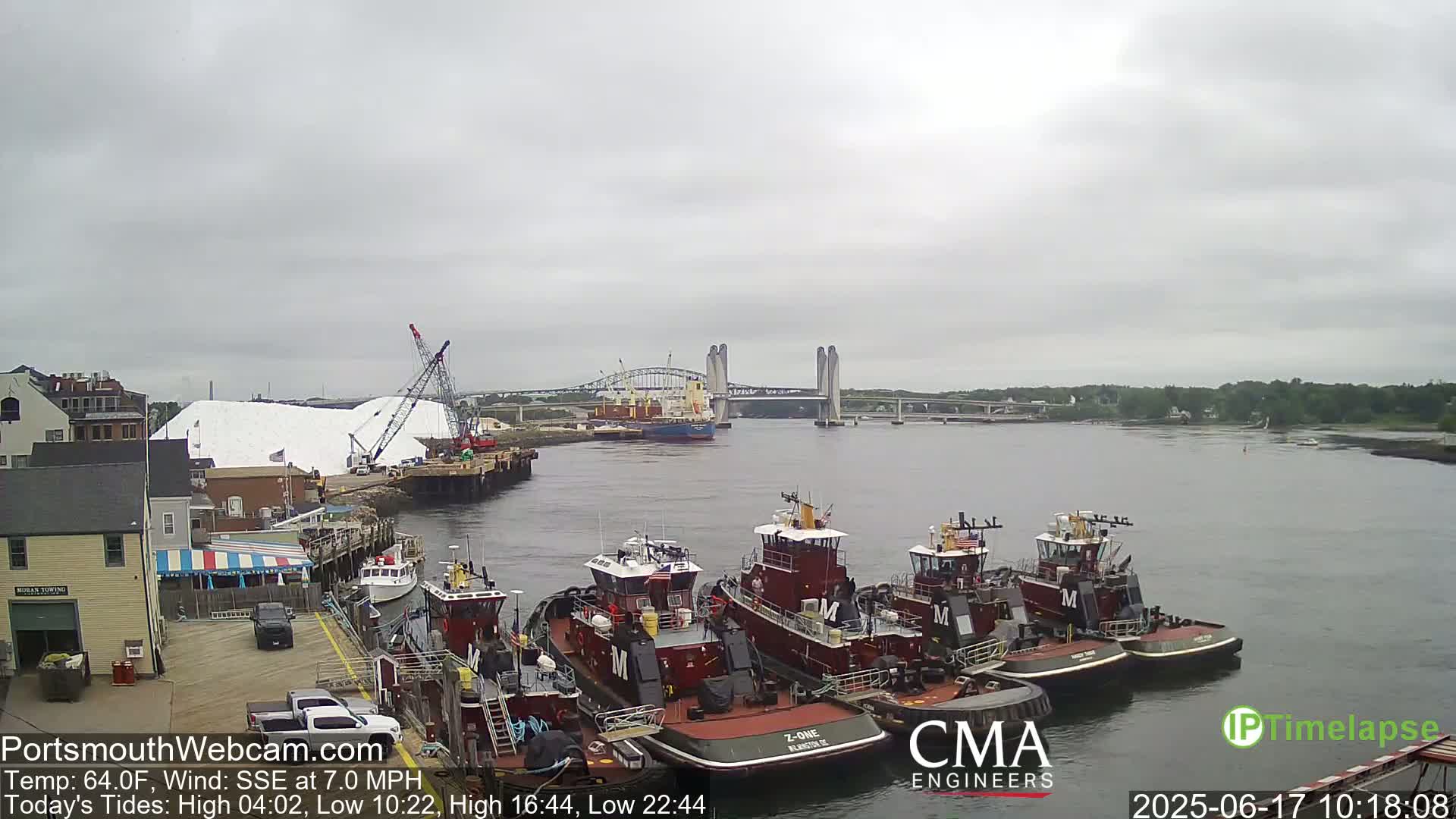 Several tugboats are moored at a dock next to a large pile of white material, with a bridge and a cargo ship visible in the distance under an overcast sky.