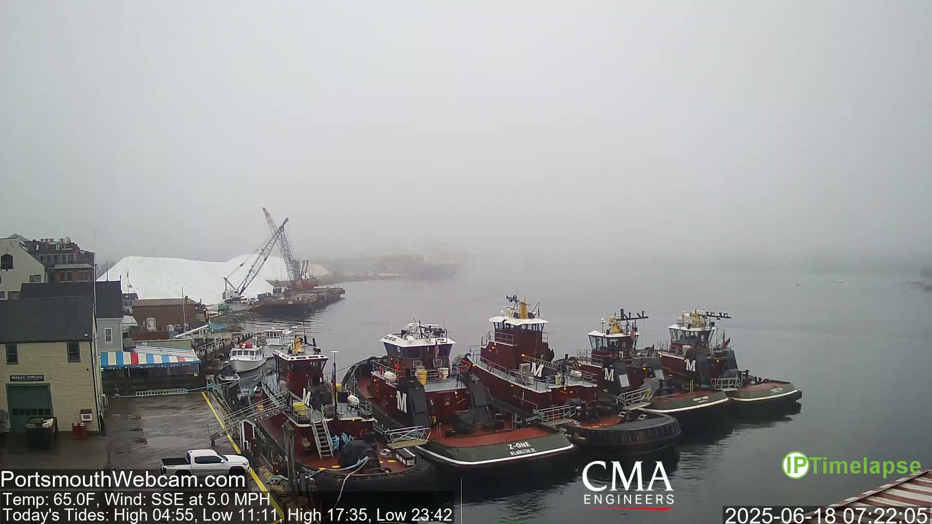 Several tugboats are moored at a dock in foggy conditions, near a large pile of what appears to be salt, which is being loaded by cranes.