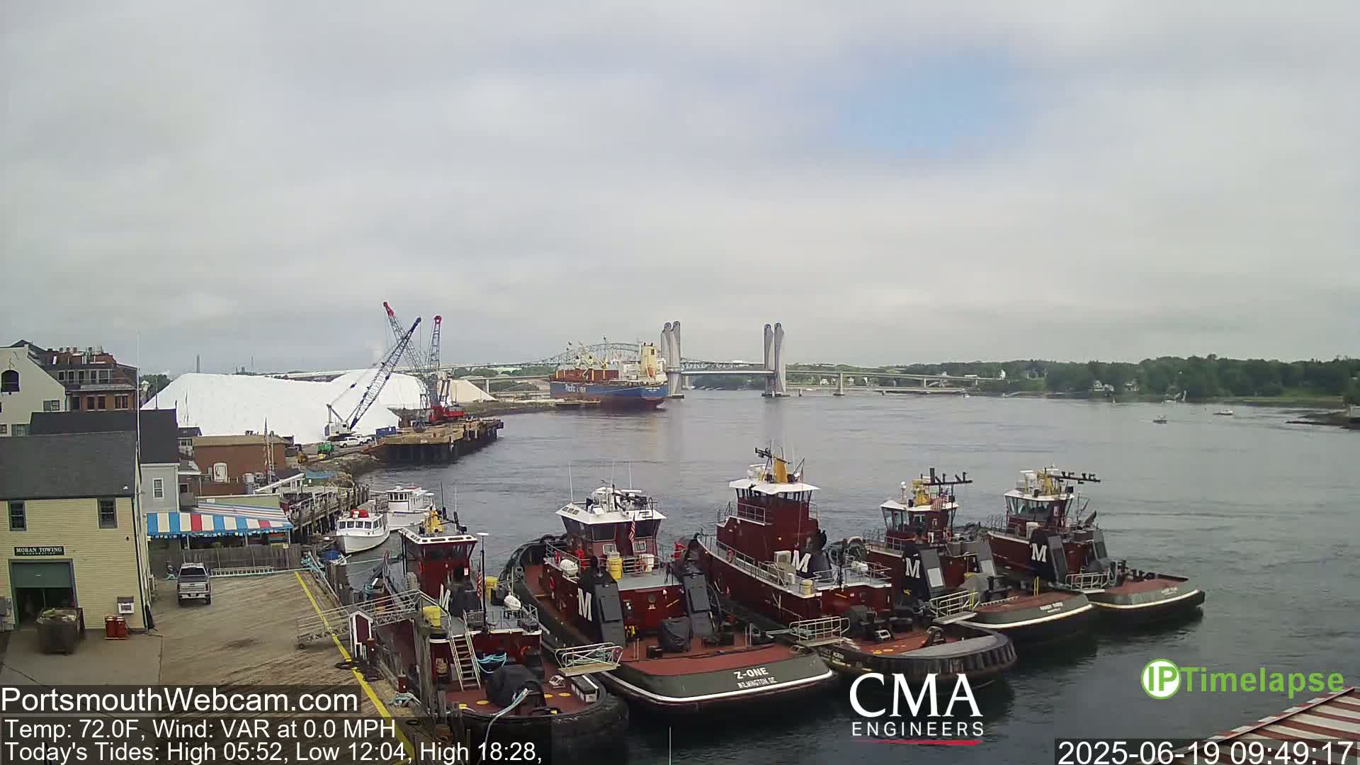A harbor scene on an overcast day shows a large pile of white material being loaded onto a ship, several tugboats moored at a dock, and a bridge in the distance.