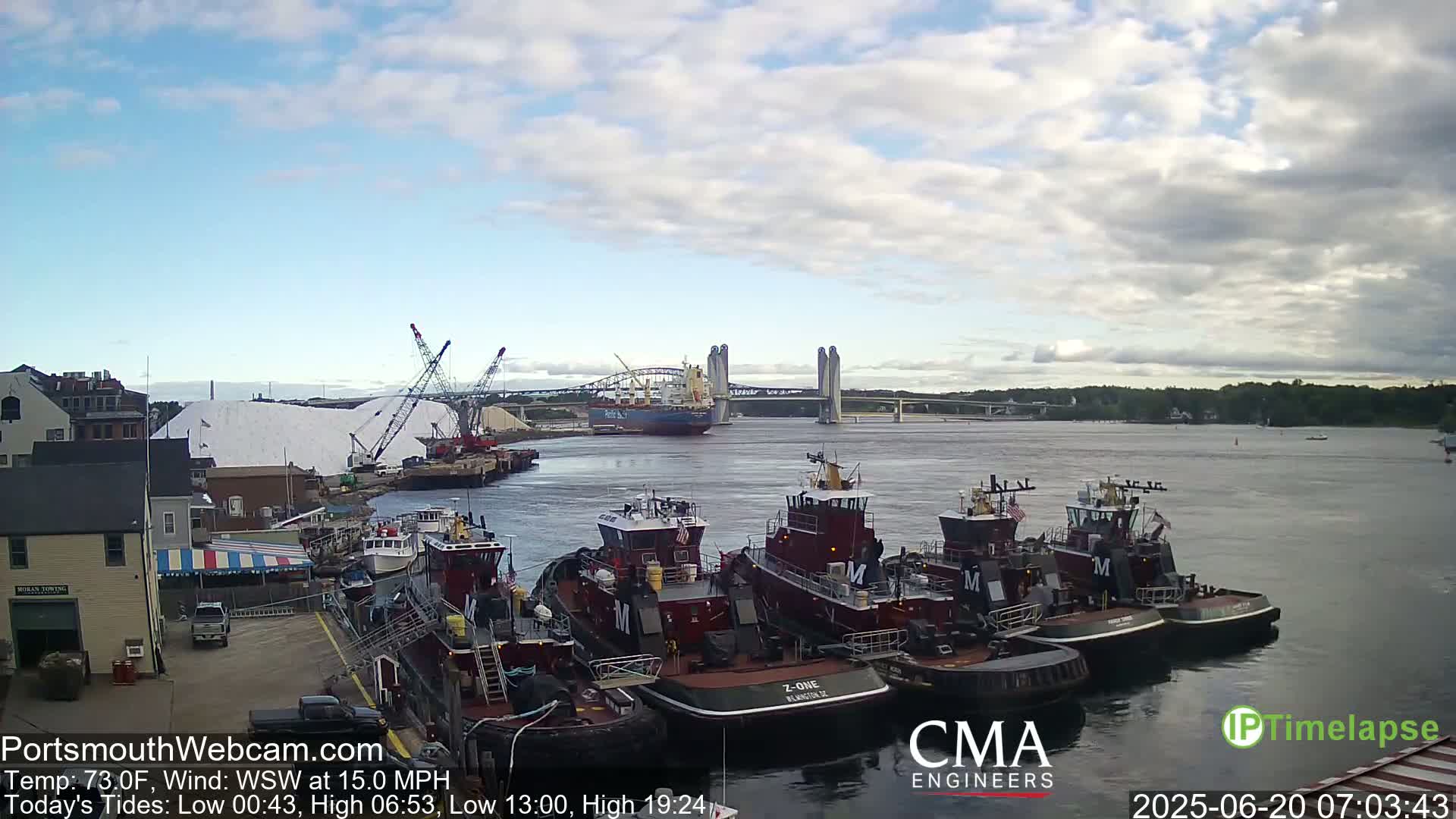 A partly cloudy day shows several tugboats docked near a large pile of white material, with a large ship and a bridge in the background.