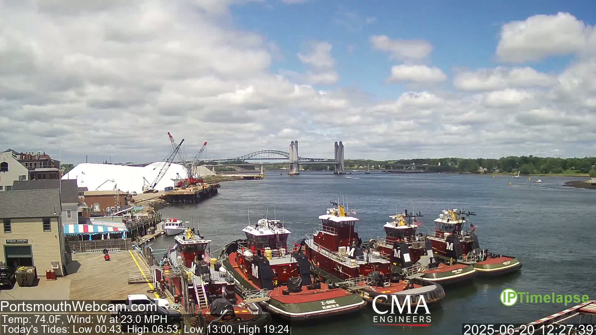 Several tugboats are moored at a dock near a bridge under a partly cloudy sky.