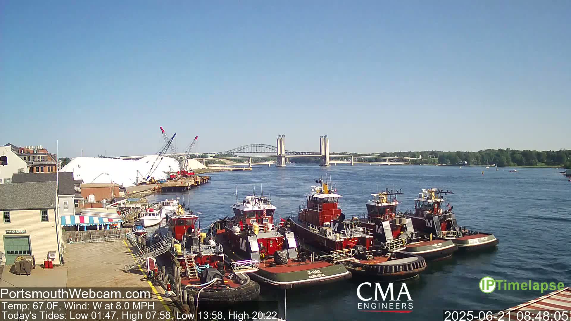 A harbor scene on a sunny day with a calm body of water shows several tugboats docked near a large pile of white material, with a bridge in the distance.