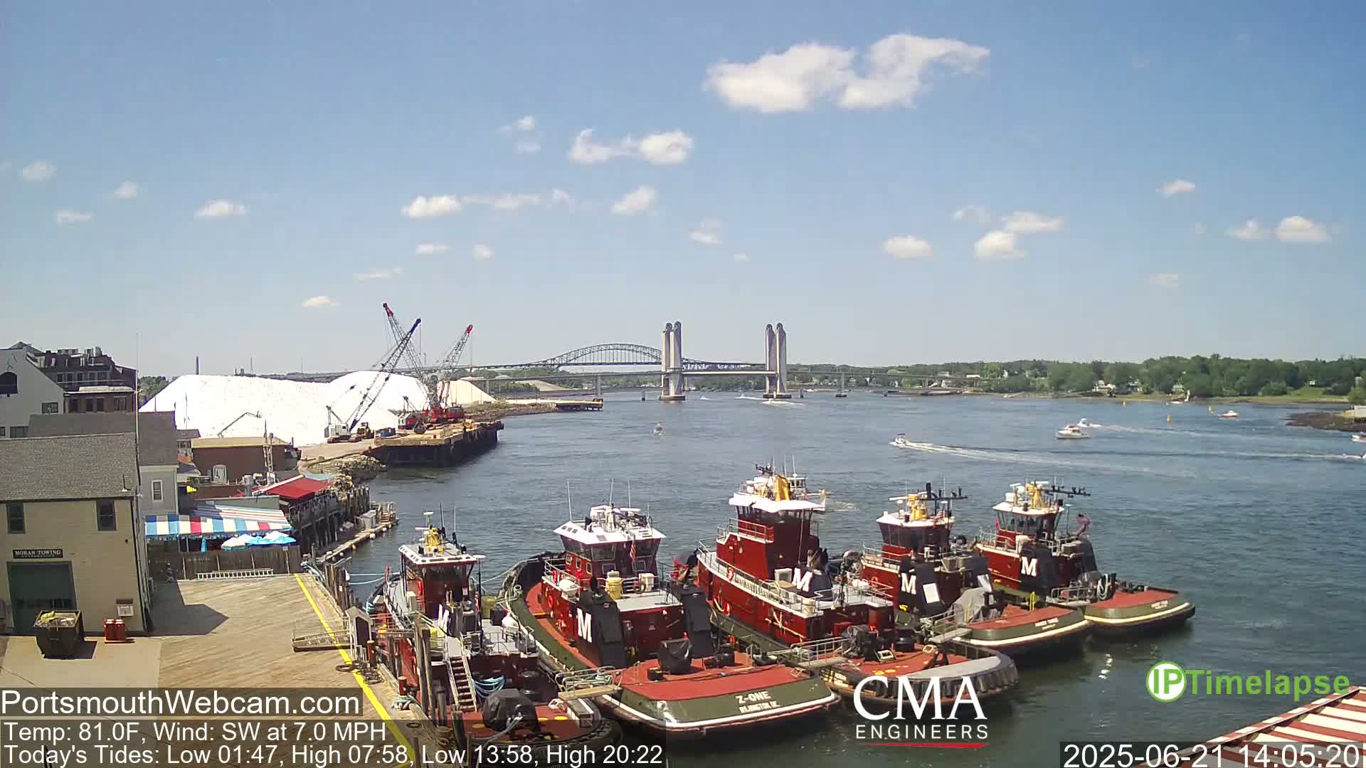 Several tugboats are moored at a dock near a bridge on a sunny day with a light southwest wind.