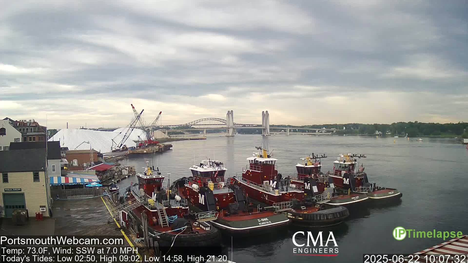 Several tugboats are moored at a dock near a bridge under a cloudy sky.
