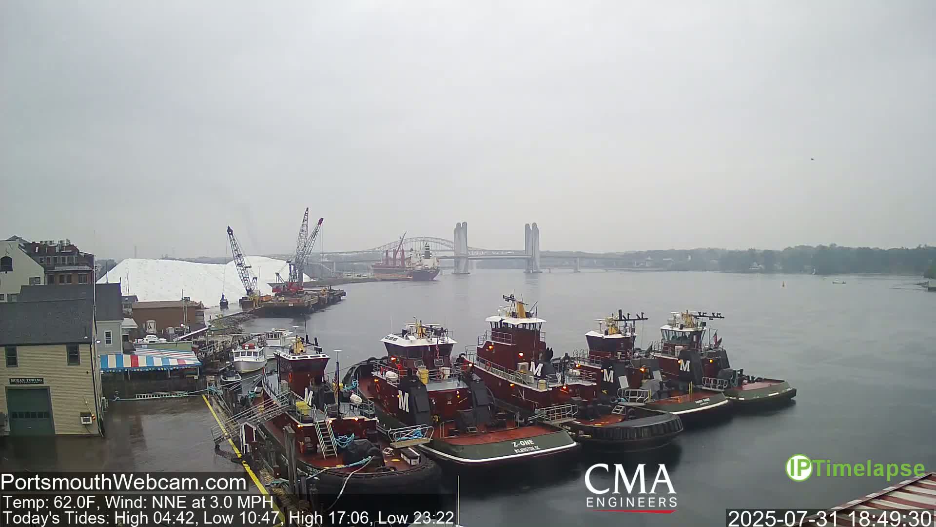 Several tugboats are docked near a large pile of white material, with a bridge and a cargo ship visible in the distance under an overcast sky.