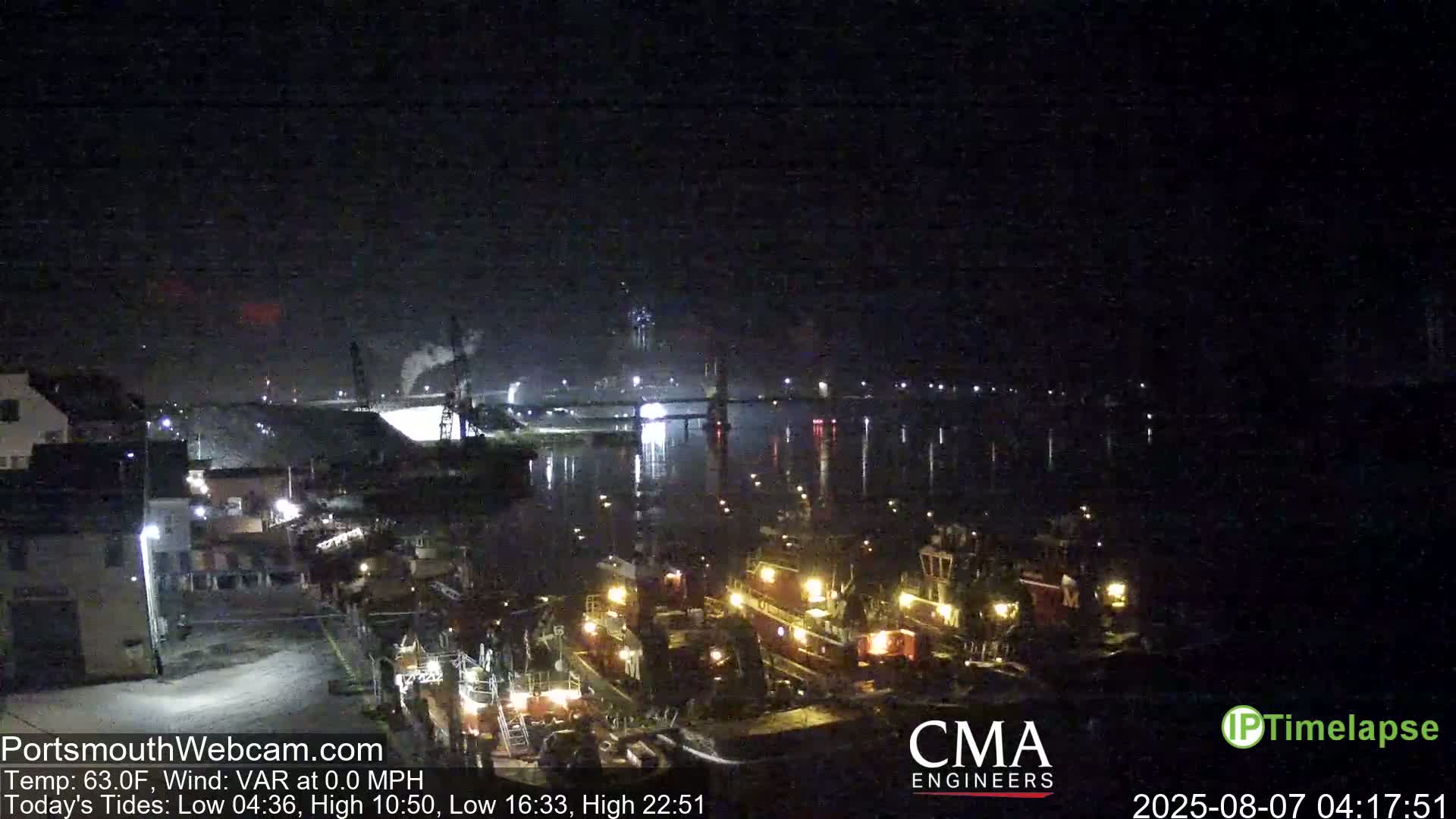 A nighttime view of a harbor with several tugboats docked, a bridge in the distance, and calm water under a dark sky.