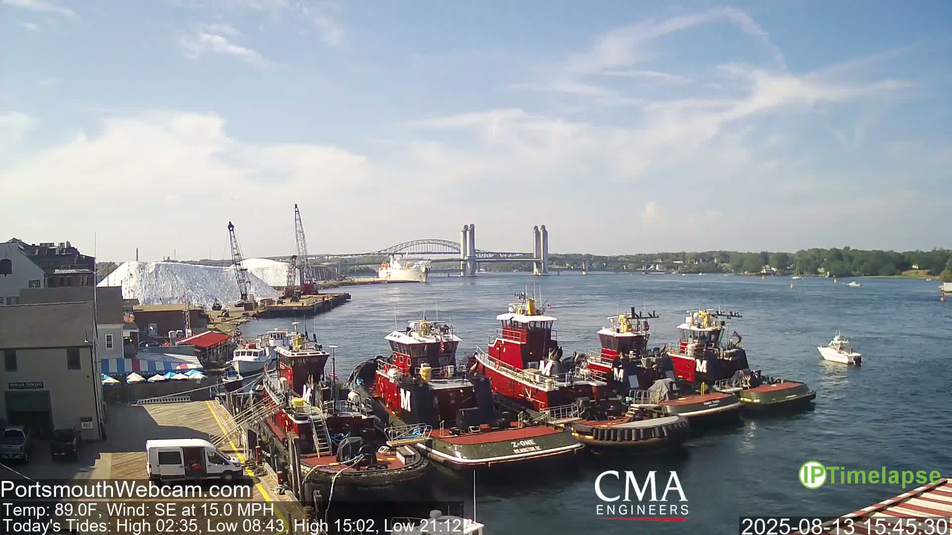 Several red tugboats are docked near a large pile of white material, with a bridge and calm, sunny water in the background.