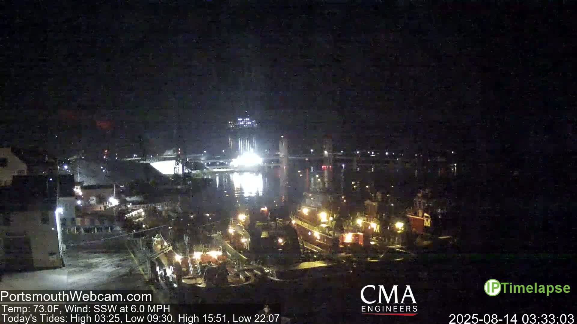 A nighttime view of a harbor with several docked tugboats, brightly lit industrial structures across the water, and a calm body of water under a clear, dark sky.