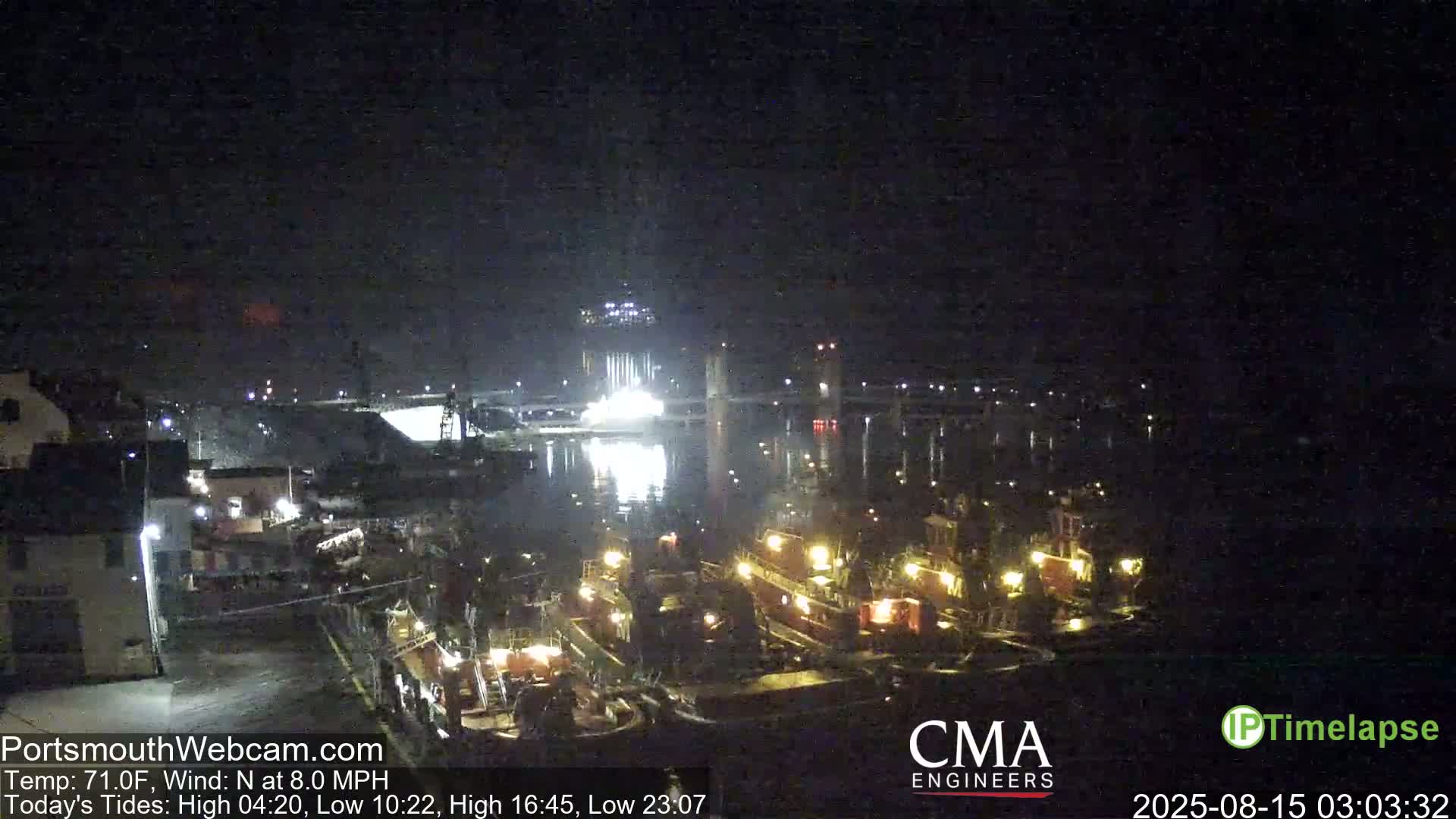 A nighttime view of a harbor with several tugboats docked, illuminated lights reflecting on the water under a dark, clear sky with a moderate north wind.
