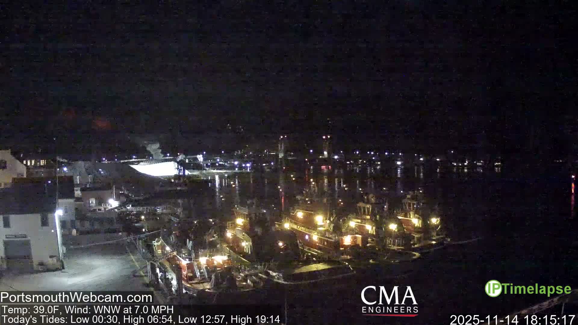 On a clear night, a harbor scene unfolds with numerous brightly lit boats docked along the waterfront, flanked by illuminated buildings and a prominent, lit bridge in the distance.