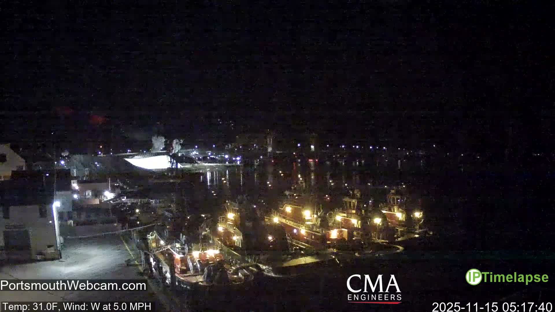 The image depicts a clear, dark night over a harbor or waterfront, with several illuminated boats docked and their lights, along with distant shoreline lights, reflecting brightly on the water, at a brisk 31.0F with a 5.0 MPH west wind.