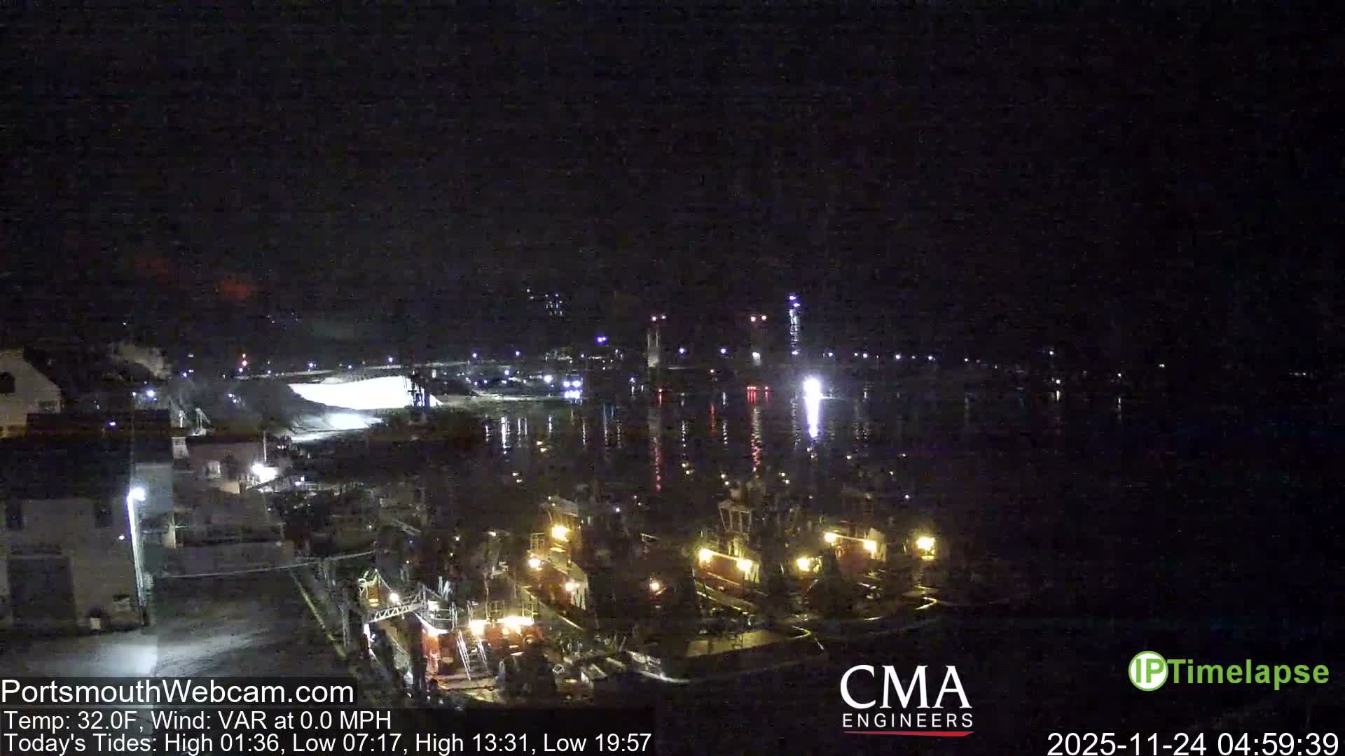 On a calm, dark night, numerous illuminated boats are docked in a harbor, their bright lights reflecting on the water, with distant bridge lights visible across the bay.