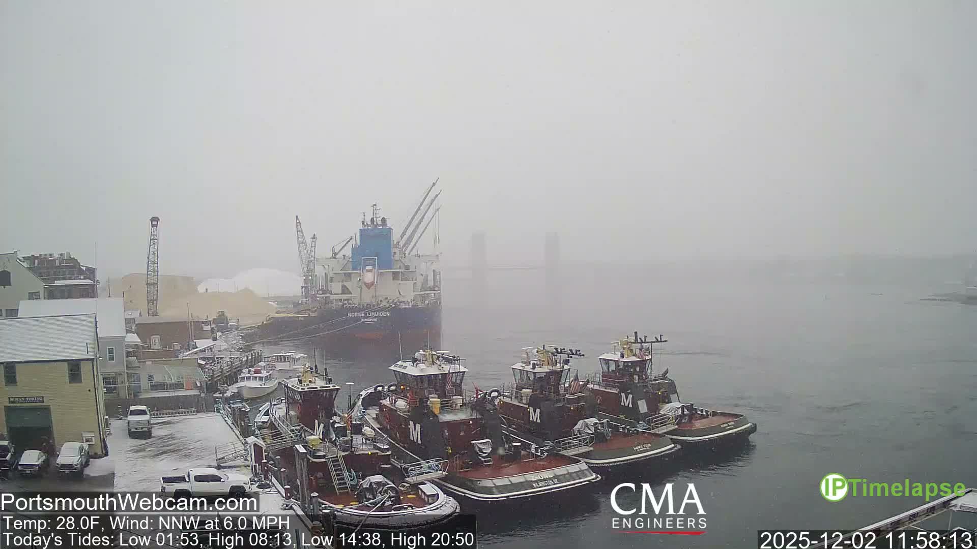 On an overcast and snowy day, a harbor scene unfolds with multiple tugboats docked in the foreground, a large cargo ship moored by docks piled with material, and buildings and distant bridge structures visible through the hazy conditions.