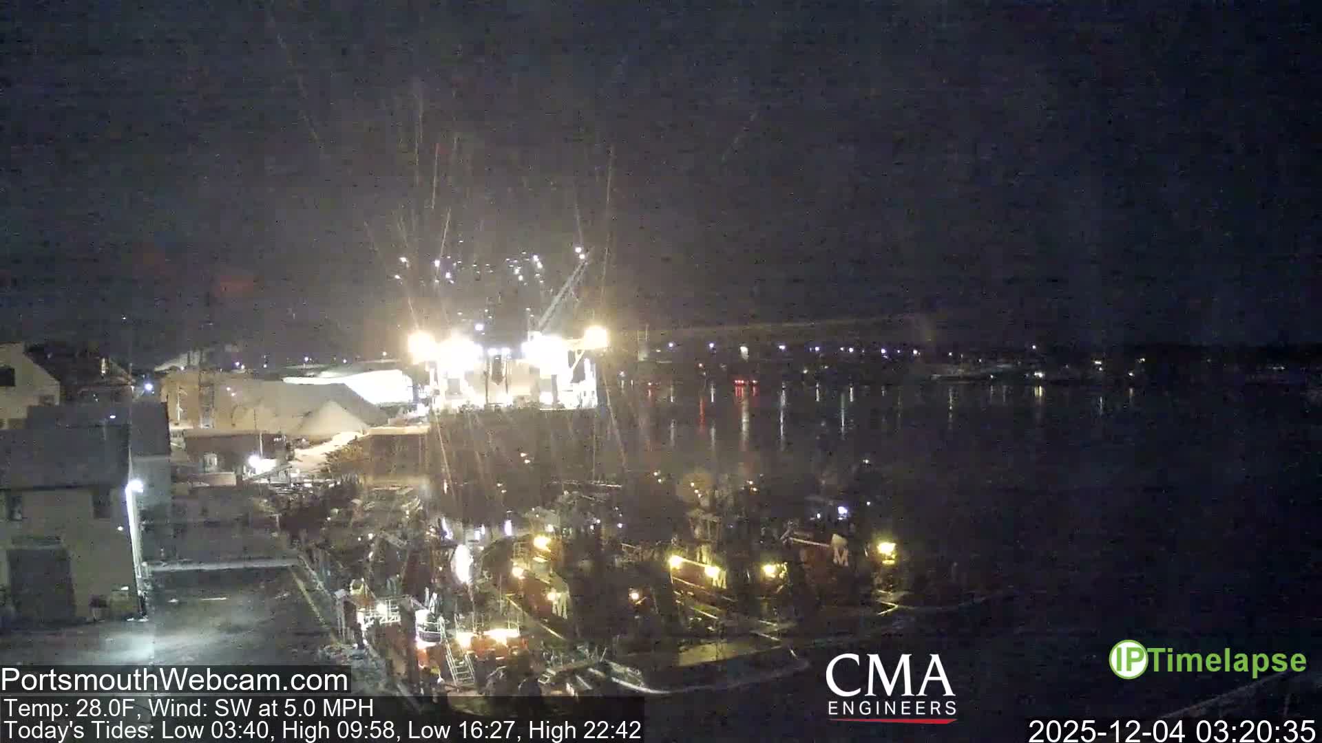 A bustling harbor at night is illuminated by numerous lights reflecting on the water, showing several boats docked and industrial structures on the shore, all under conditions of falling rain or snow.