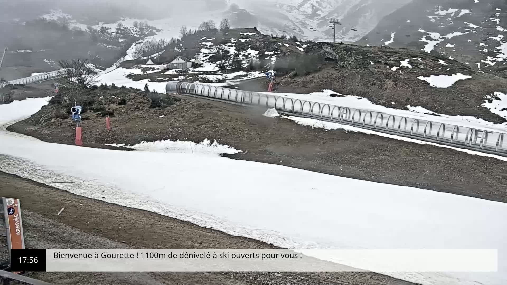A snowmaking machine operates on a ski slope in overcast weather, with a long, covered run and a partially snow-covered ground visible.