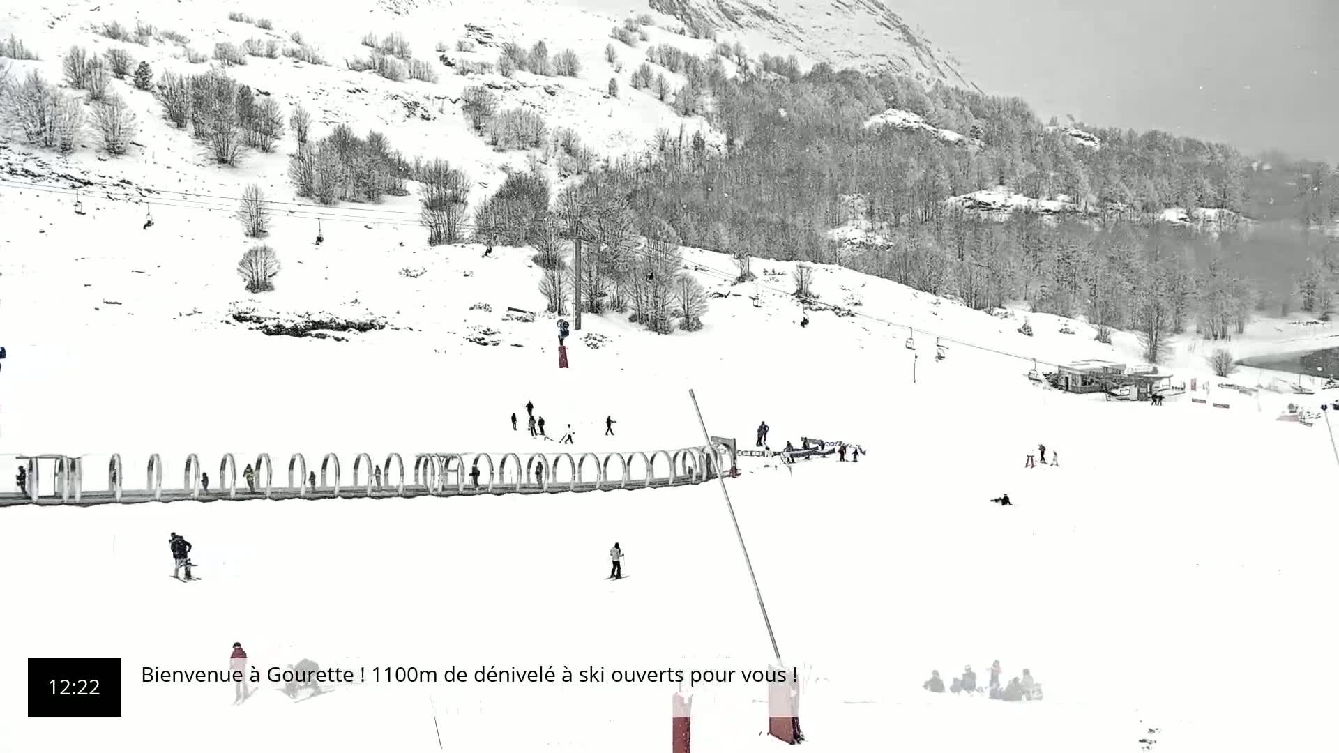 A snowy ski slope with several skiers and snowboarders, a ski lift, and a small building, under a light snowfall.