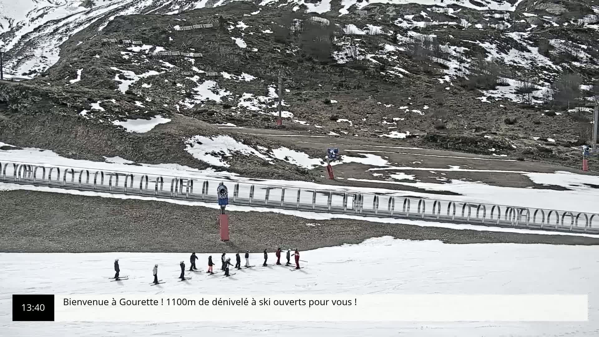 A group of skiers stands on a snowy slope at the bottom of a rocky, partially snow-covered mountain.