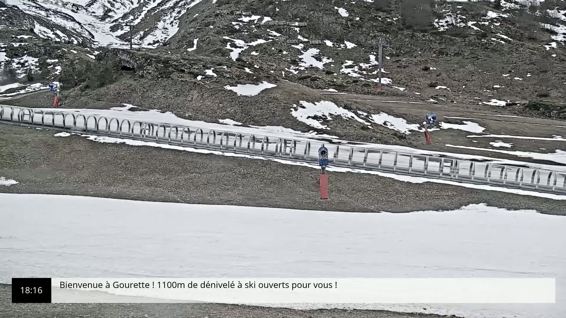 A snow-covered mountain slope with a metal railing and patches of bare ground, under a mostly clear sky.
