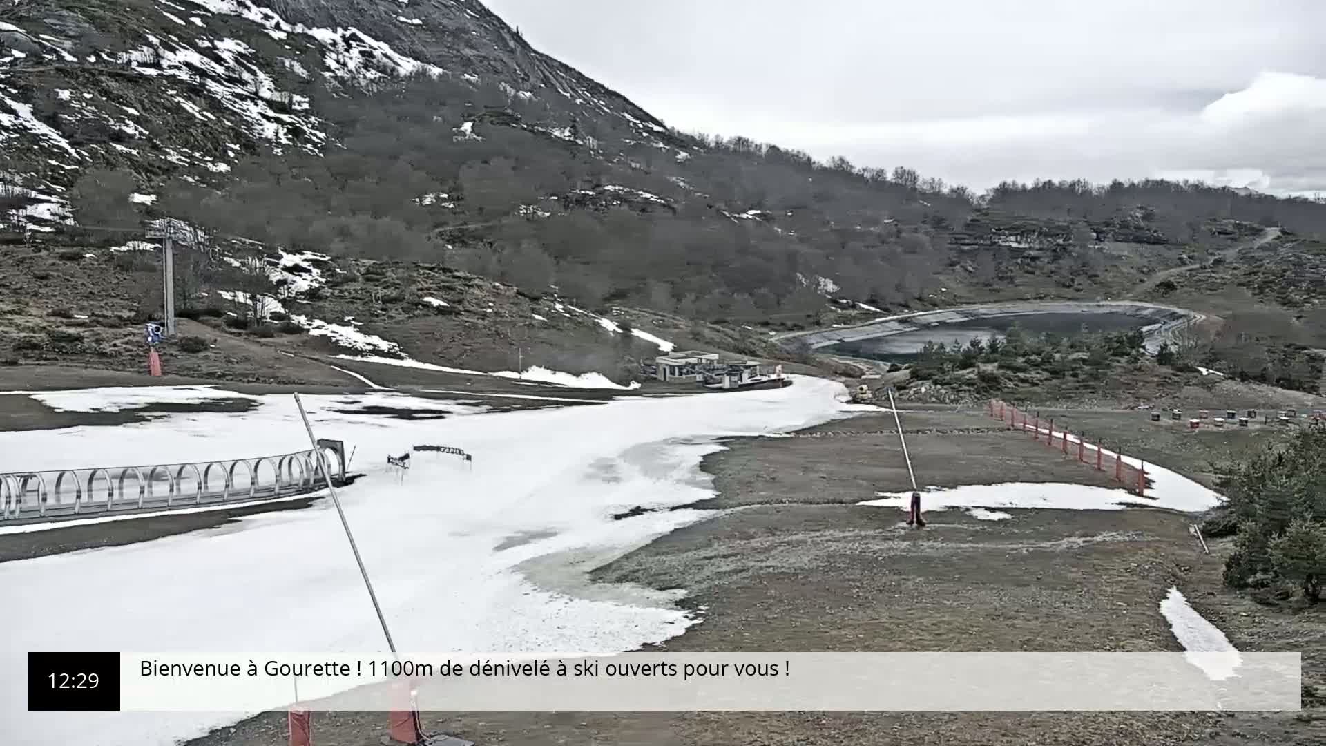 A mostly cloudy day shows a ski slope with patches of melting snow and ice on a mountainside, near a small body of water.