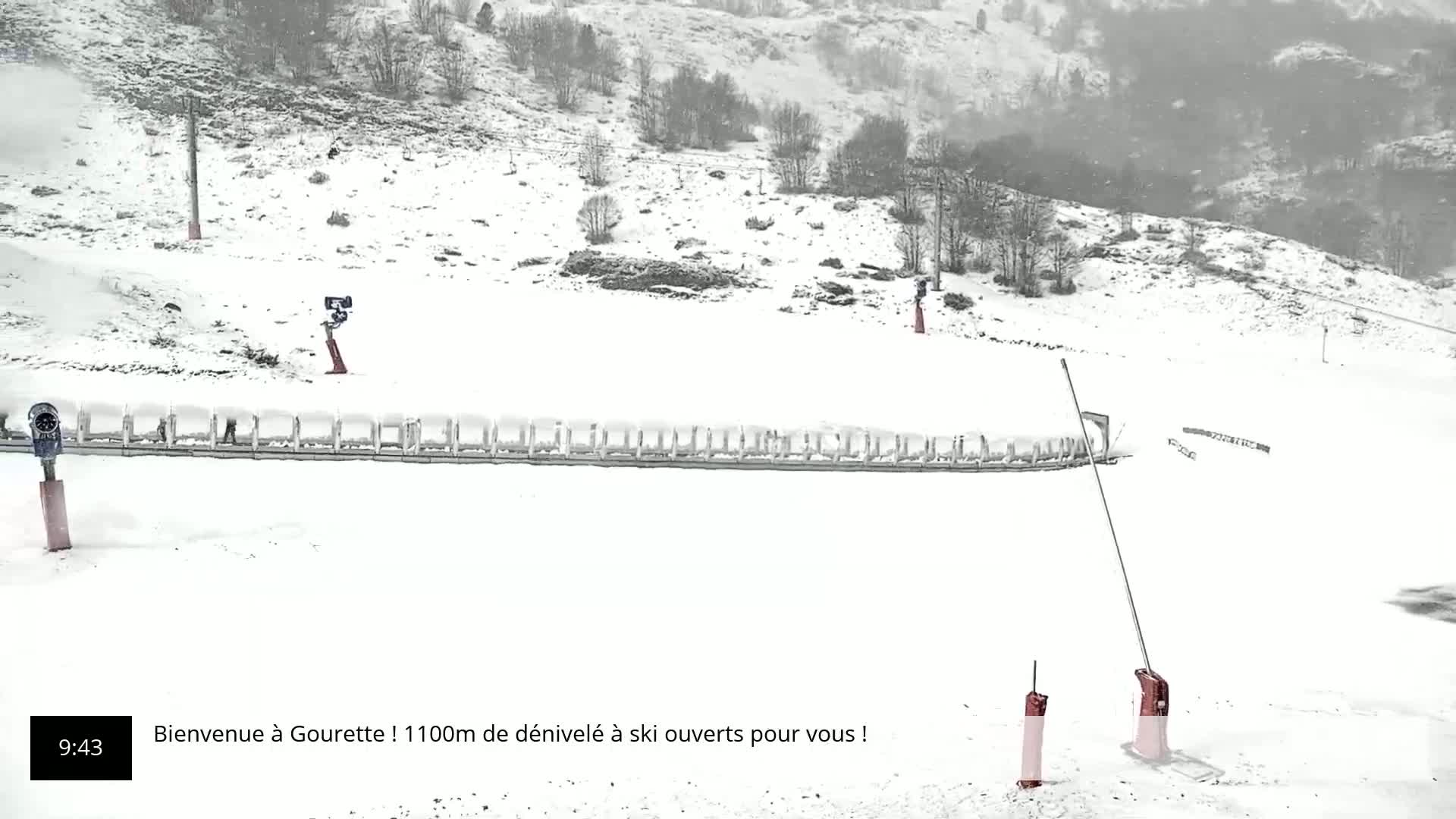 A snow-covered ski slope with several snowmaking machines and ski lift equipment is visible under heavy snowfall.