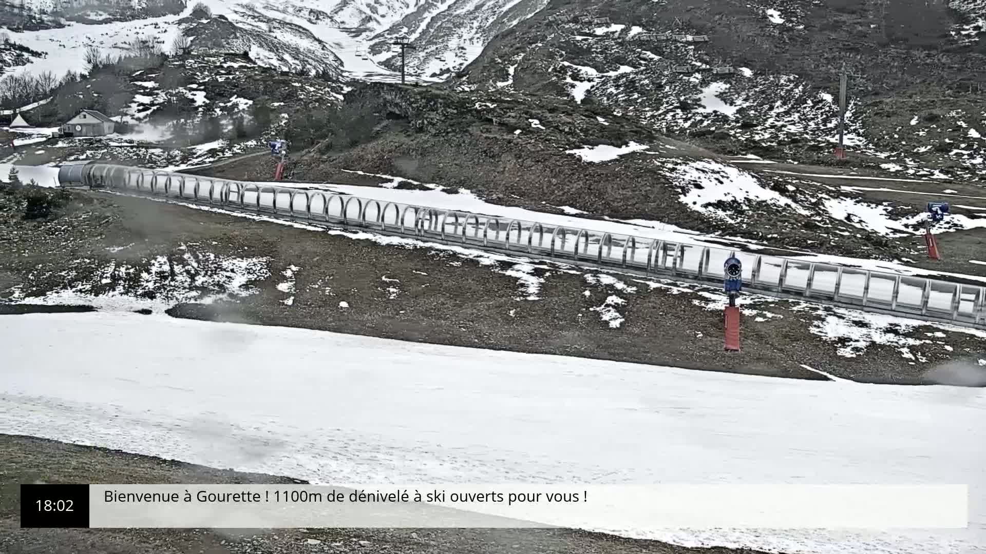 A long, covered ski lift extends across a snow-covered slope in a mountainous area, with snowmaking machines visible and some patchy snow on the ground.