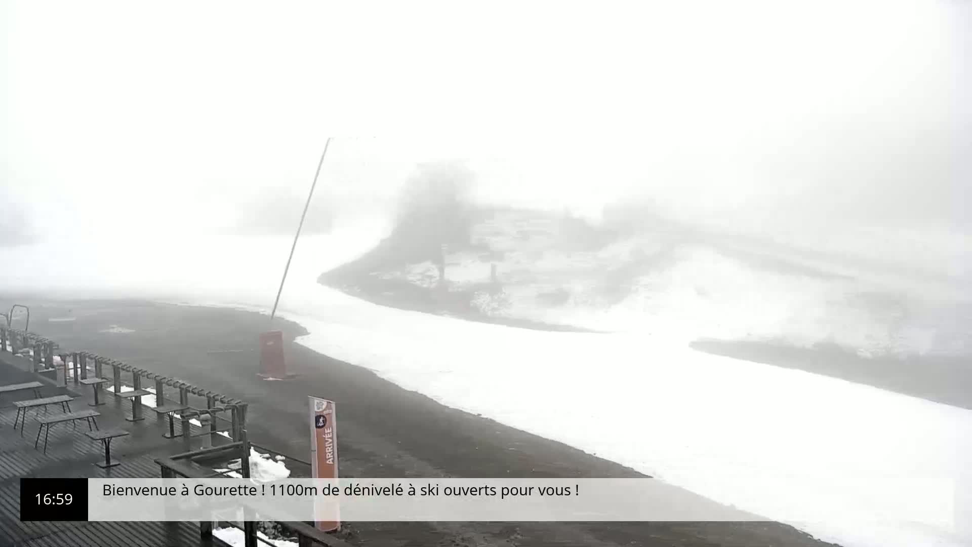 A snow-covered ski area is shrouded in thick fog, with a paved area and outdoor furniture visible in the foreground.