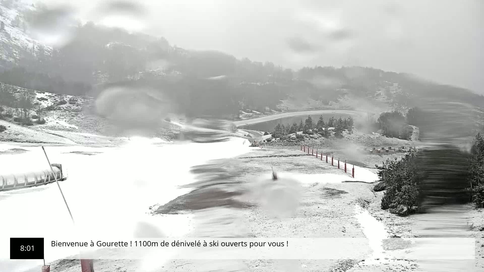 A snowy, overcast scene shows a ski slope with a snowmaking machine operating in the foreground, and a mostly snow-covered mountainside in the background.