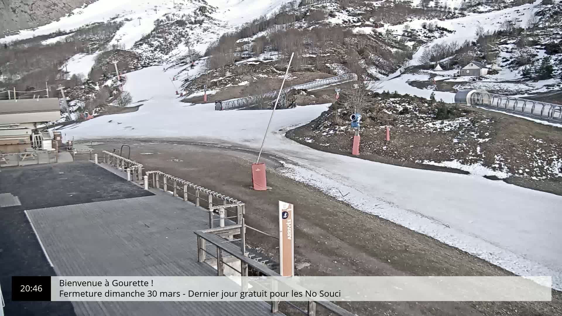 A snow-covered ski slope with a base area showing patches of bare ground and ice, under a cloudy sky.