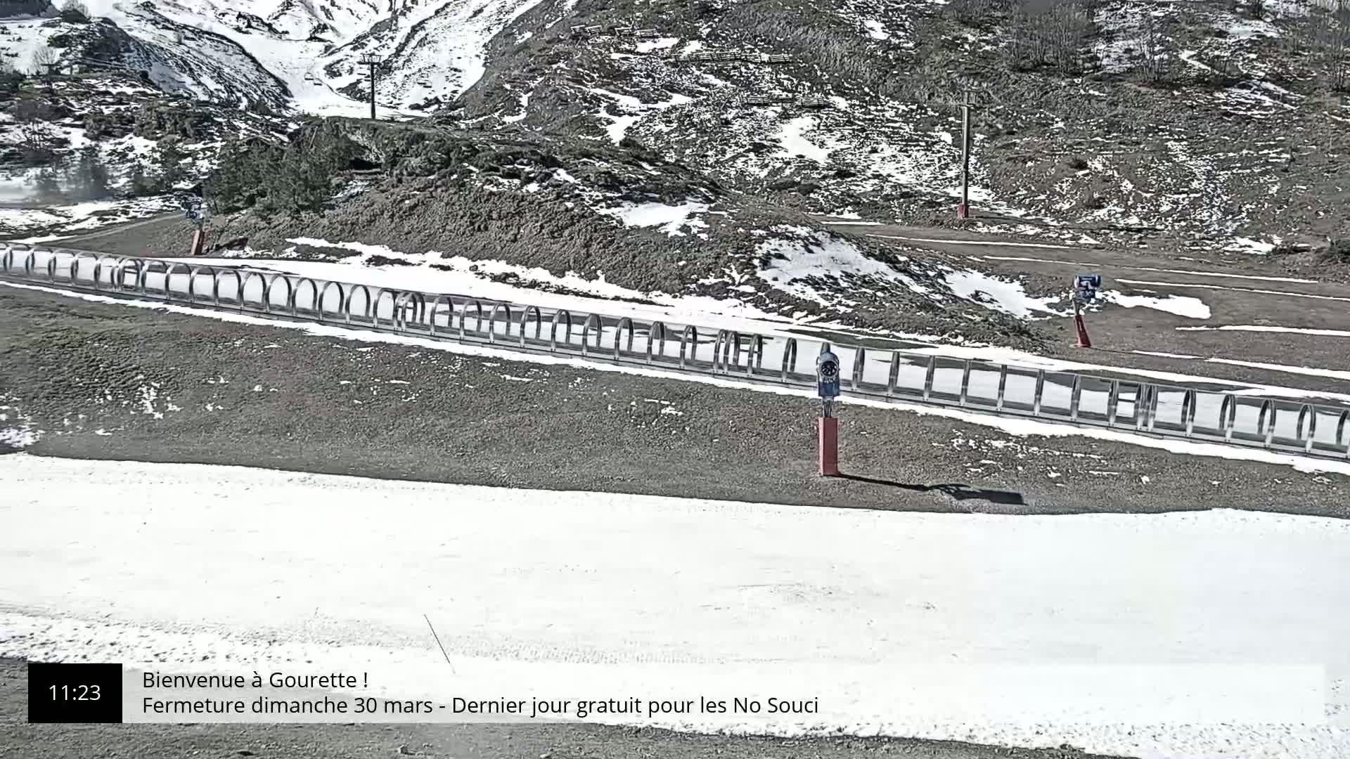 A snowy mountainside scene shows a paved area with a snow-covered section, partially enclosed by a metal railing, and a small weather station.
