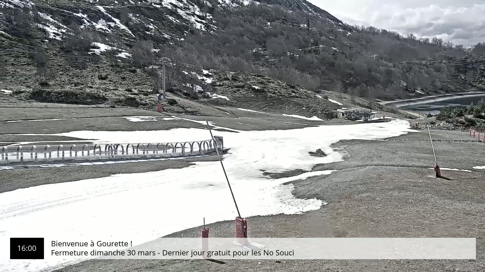 A mostly clear day shows a mountainside ski slope with patchy snow, a small building, and a calm lake in the background.