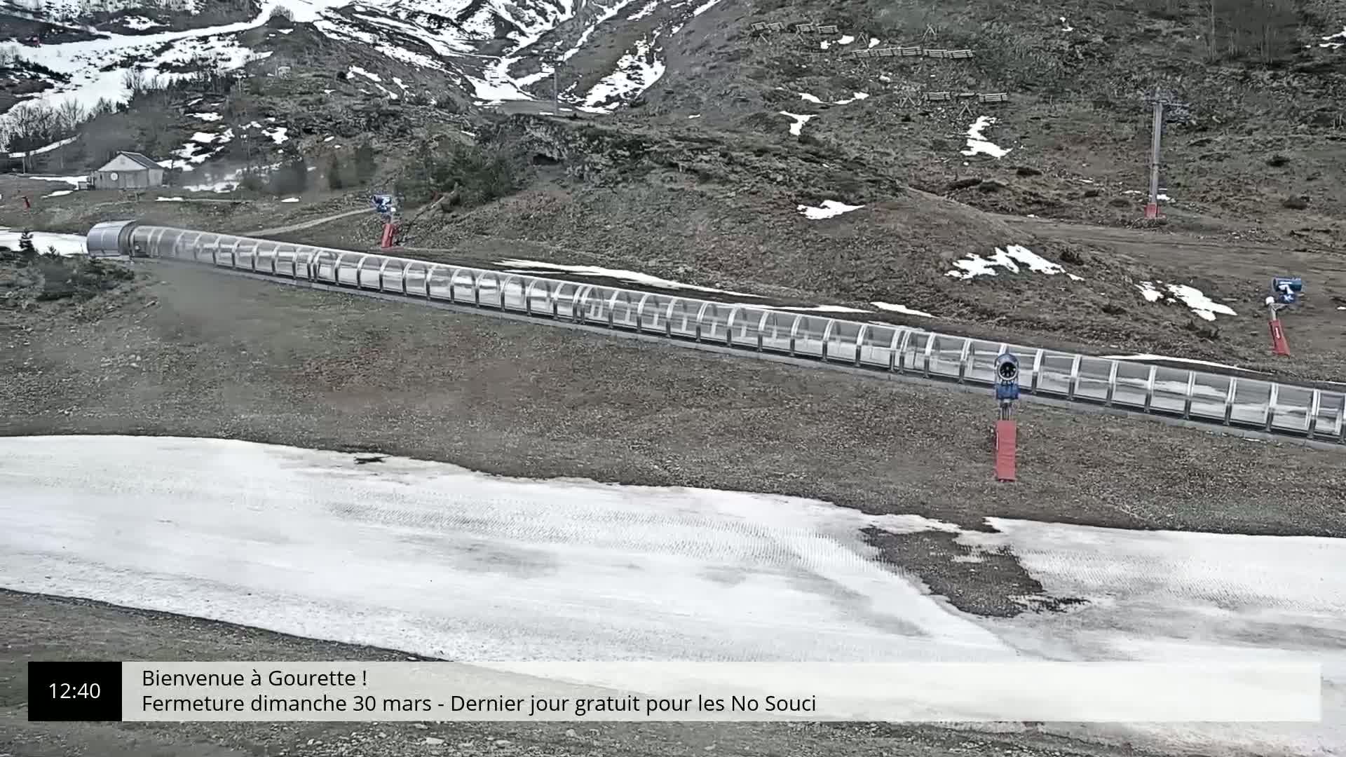 A long, covered toboggan run extends across a mostly barren, rocky landscape with patches of snow and ice, situated against a mountainous backdrop with some remaining snow.