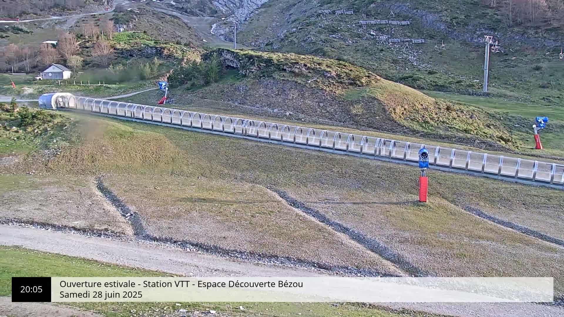A long, covered track extends downhill from a small building in a mountainous, grassy area.