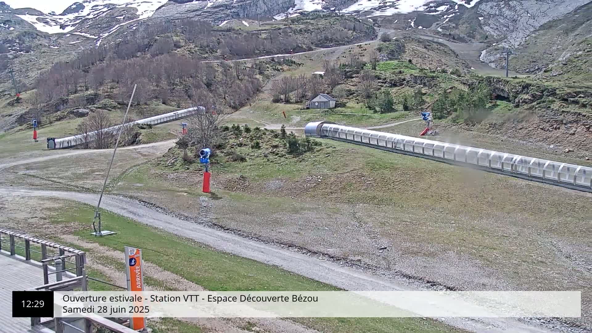 Two long covered conveyor belts are situated on a grassy, gravelly area beneath a mountain partially covered in snow, under a partly cloudy sky.