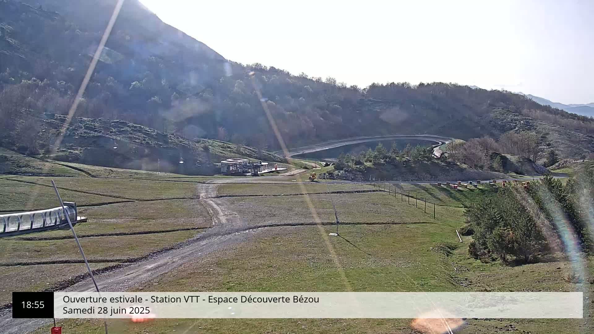 A sunny, outdoor view of a mountainous landscape shows a dirt road leading to a small building near a reservoir, with grassy fields and sparse trees.