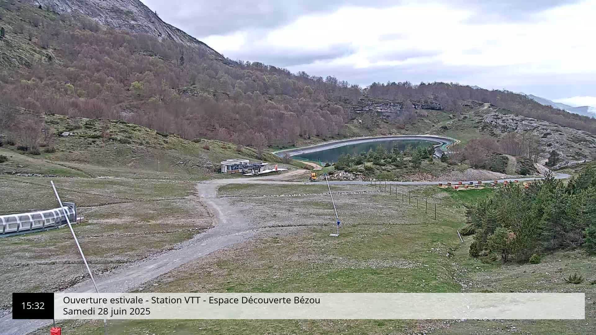 A mountaintop view shows a dirt road leading to a small building near a reservoir, under a mostly cloudy sky.