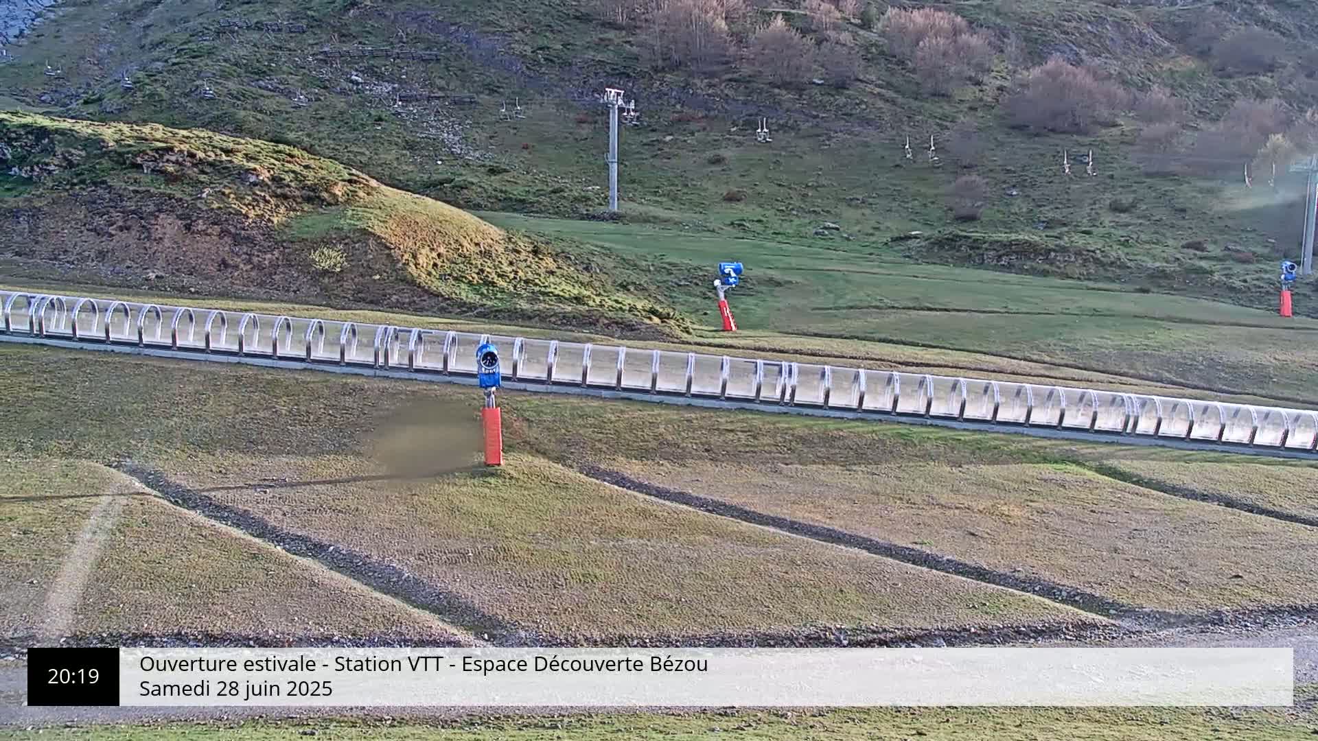 A long, covered track with two snowmaking machines sits on a grassy hillside under a sunny sky.