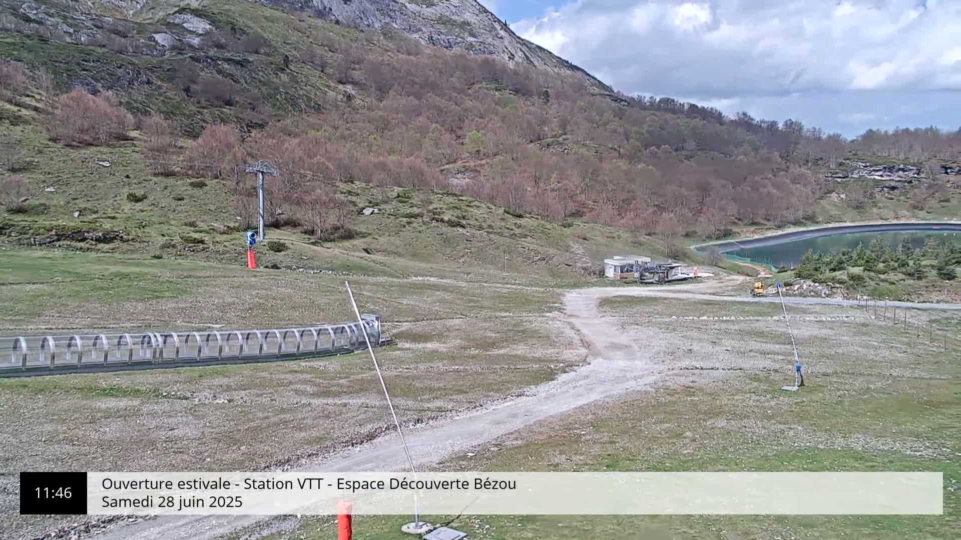 A mountainside view shows a dirt path leading to a chairlift and a covered track, under partly cloudy skies.